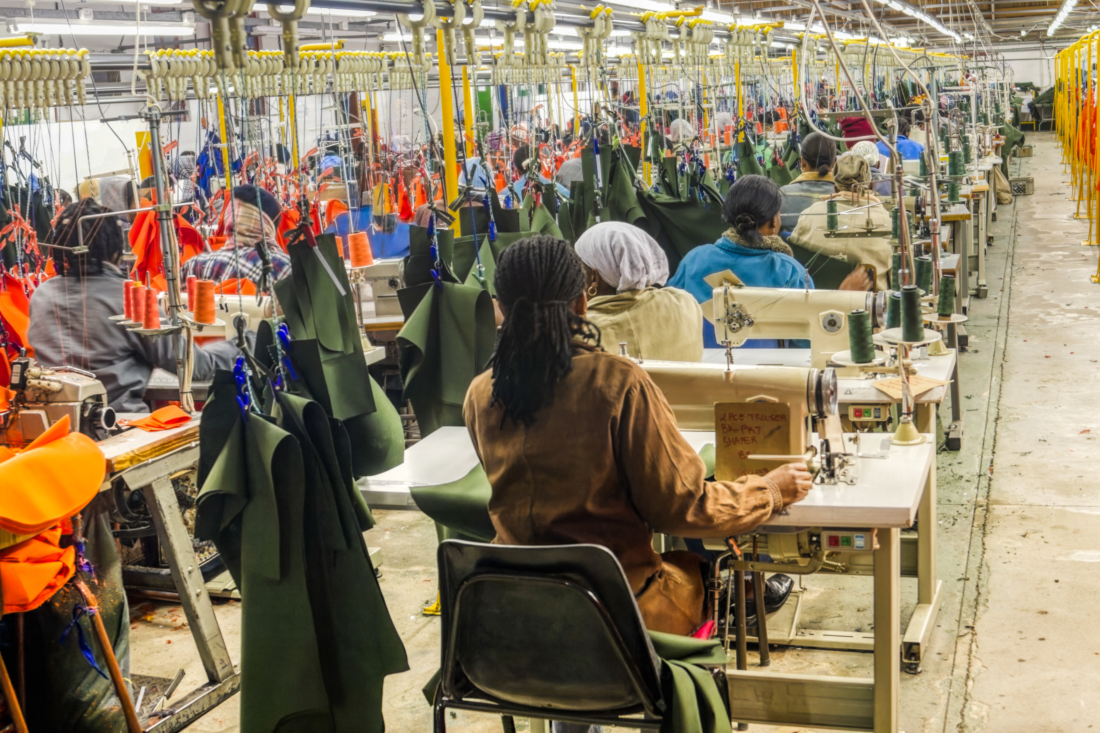 Rows of workers operate industrial sewing machines inside a large garment manufacturing factory.